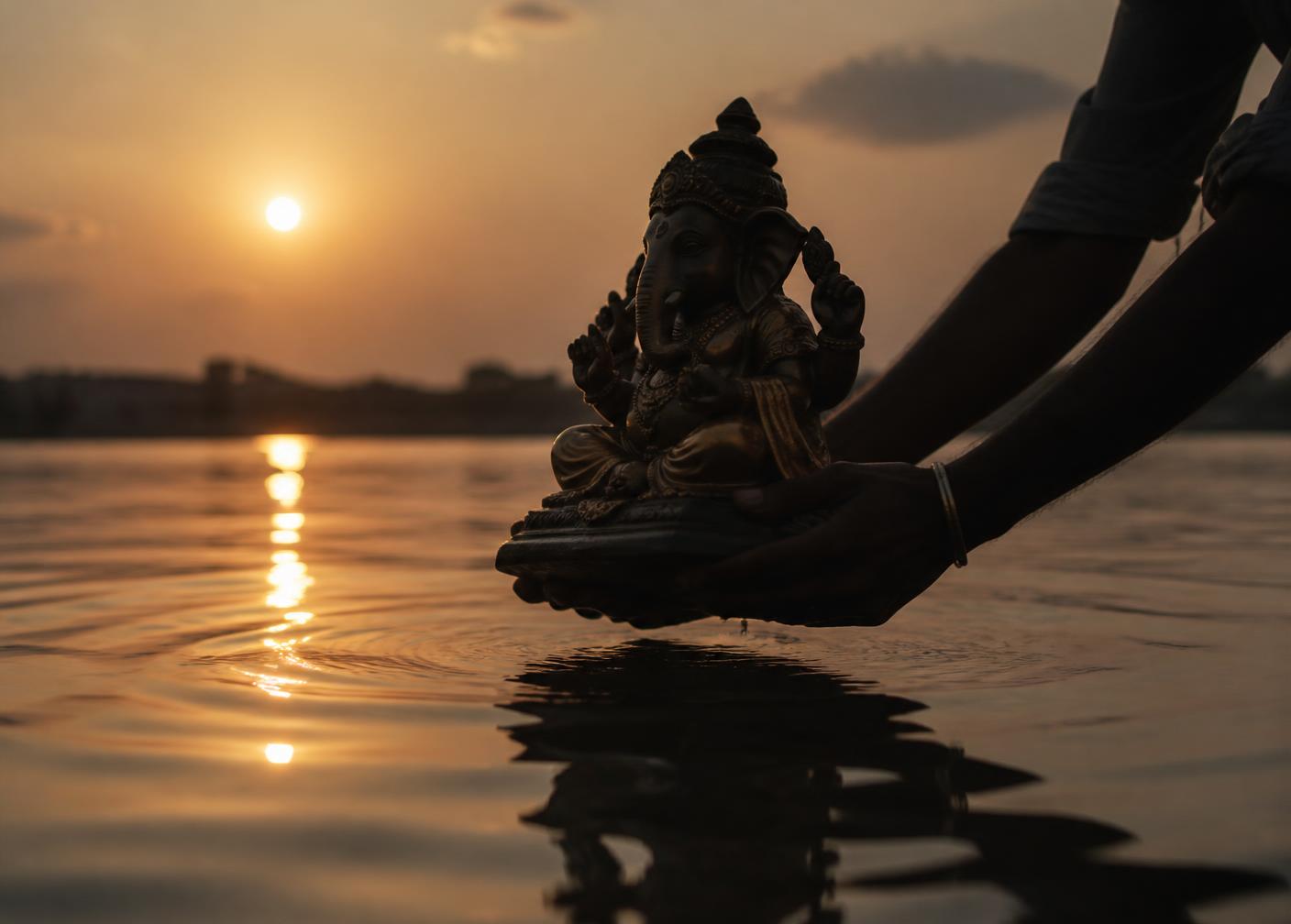 Hands gently lowering a small clay Ganesha idol into still water at dusk